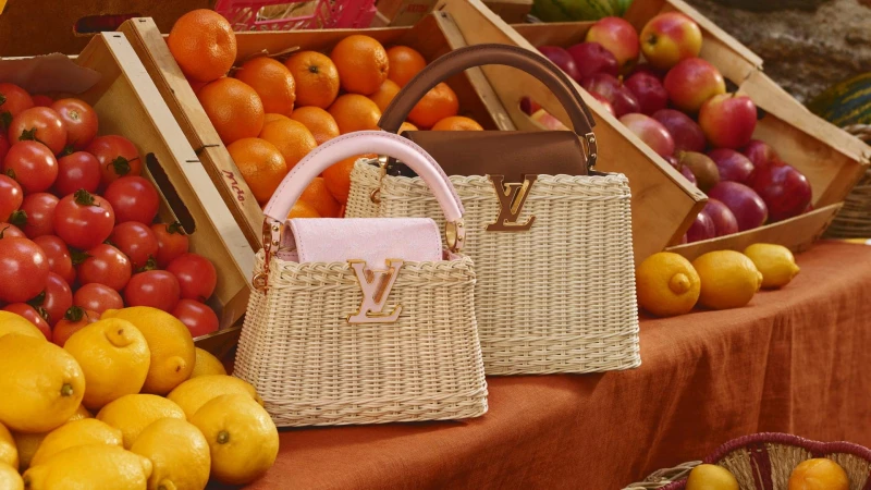 Two woven wicker handbags with leather handles and gold LV hardware displayed among crates of fresh fruit on a market table.
