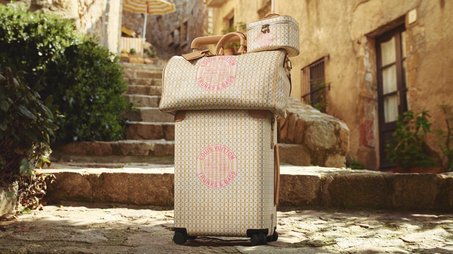 Stacked Louis Vuitton monogram luggage set on cobblestones in a European village street under warm sunlight.