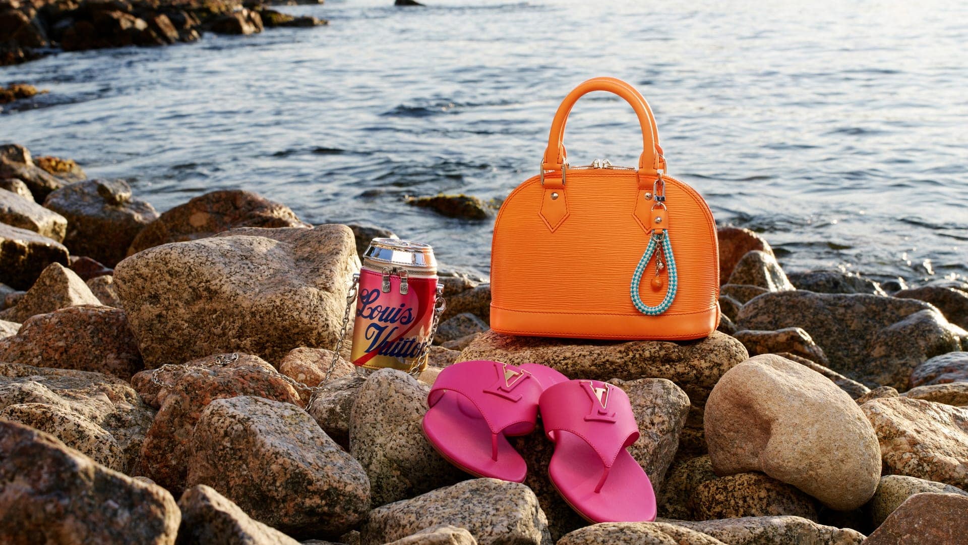 Bright orange leather handbag, pink sandals, and branded canister resting on rocky shore near ocean water.