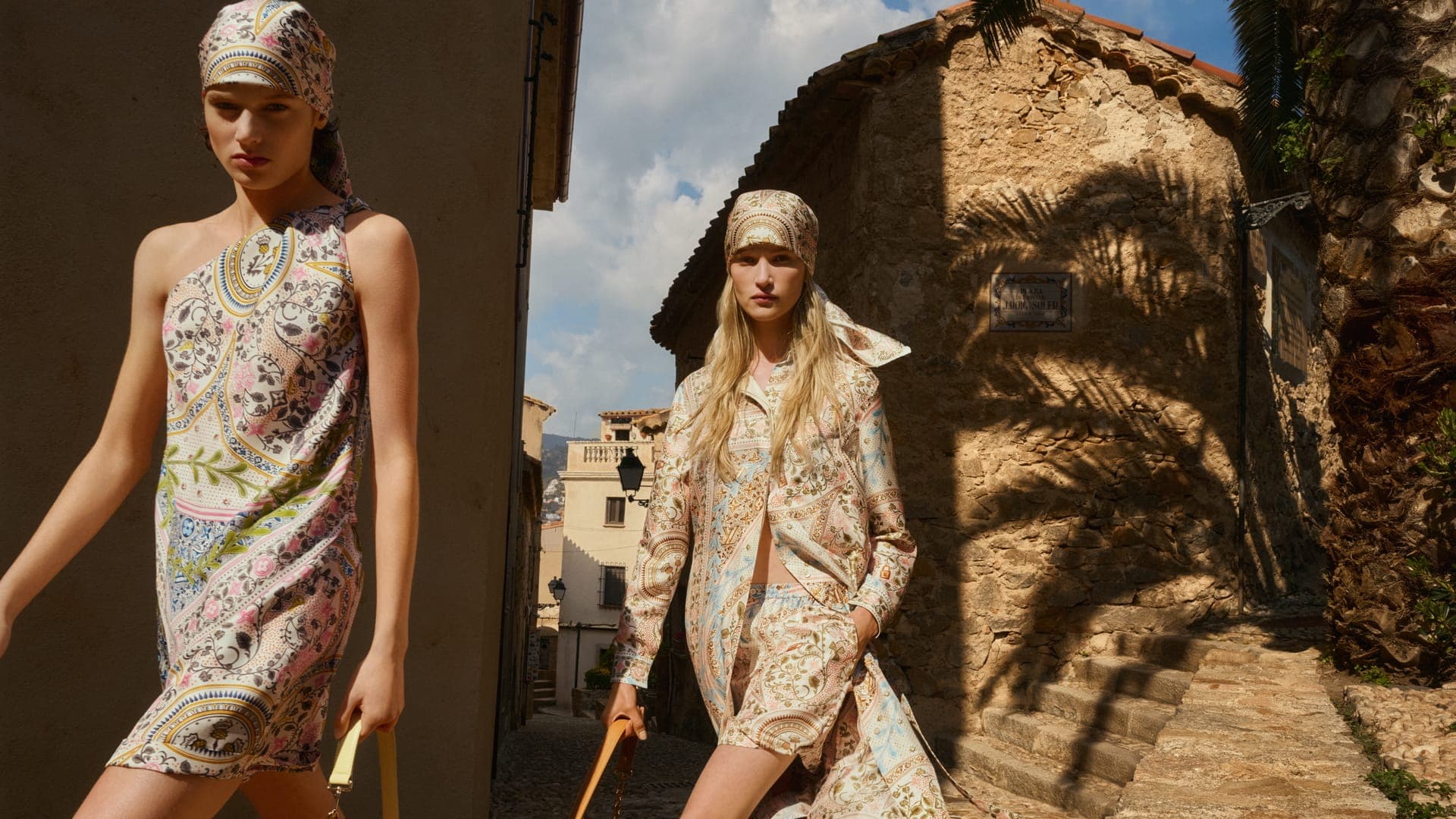Two models wearing patterned clothing and matching headscarves walking down a sunny narrow street alongside rustic stone buildings