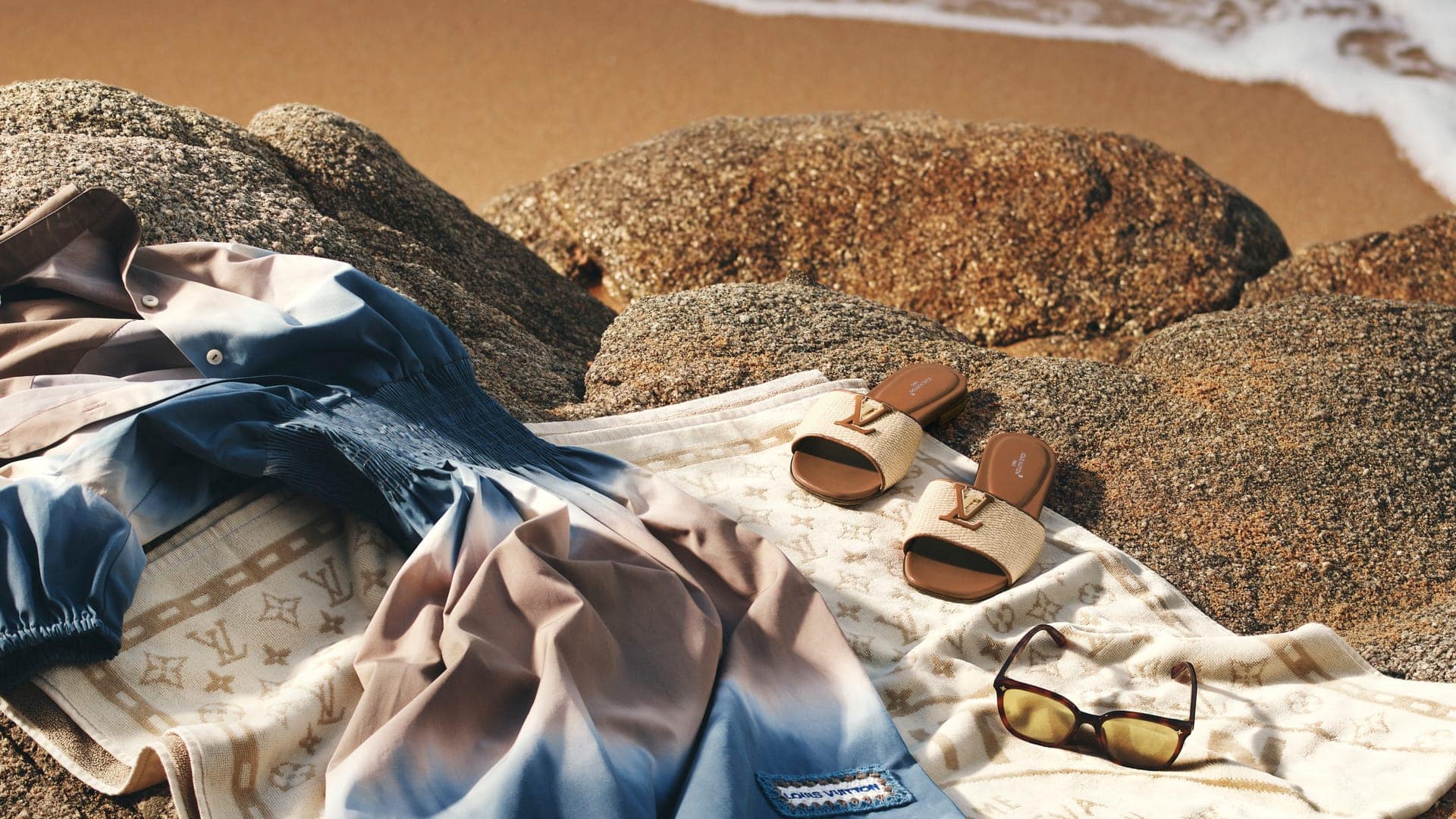 Blue ombre dress, monogram slides, and sunglasses resting on large coastal rocks by the water for Louis Vuitton Resort 2026 ad campaign.