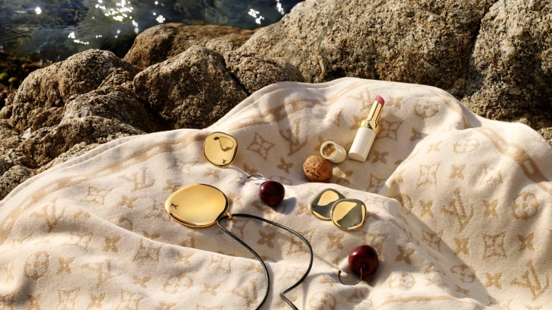 Louis Vuitton luxury accessories including gold pendant, compact, and lipstick displayed on a monogrammed blanket near rocks and water