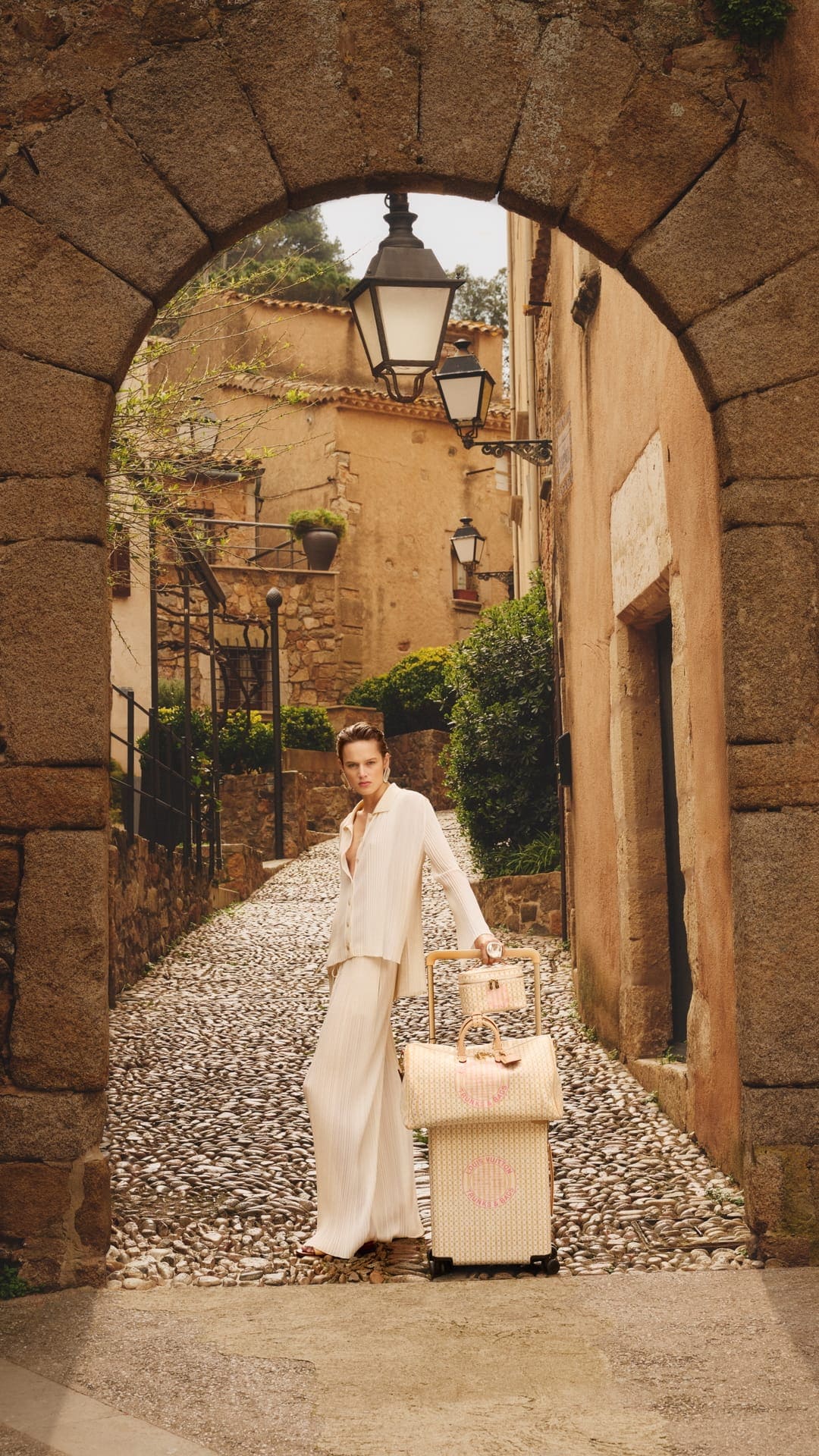 Model standing beneath a stone archway on a cobblestone street, holding luggage for Louis Vuitton Resort 2026 ad campaign.