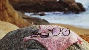 Tortoiseshell sunglasses resting on a pink patterned towel on a rock near a sandy beach and ocean waves.