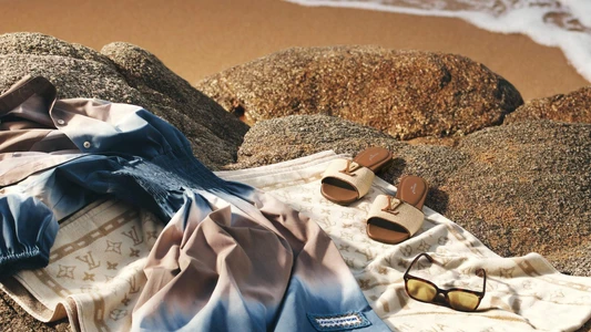 Blue ombre dress, monogram slides, and sunglasses resting on large coastal rocks by the water for Louis Vuitton Resort 2026 ad campaign.