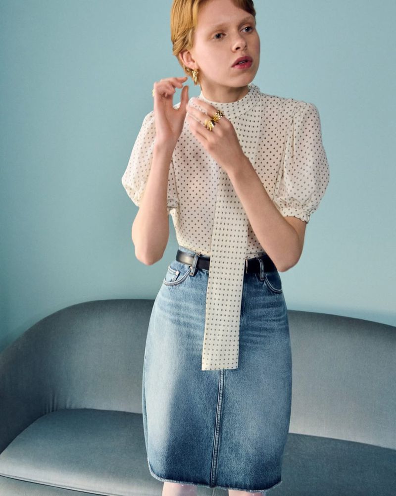Model in dotted high-neck blouse and denim skirt adjusting earrings in front of a blue sofa