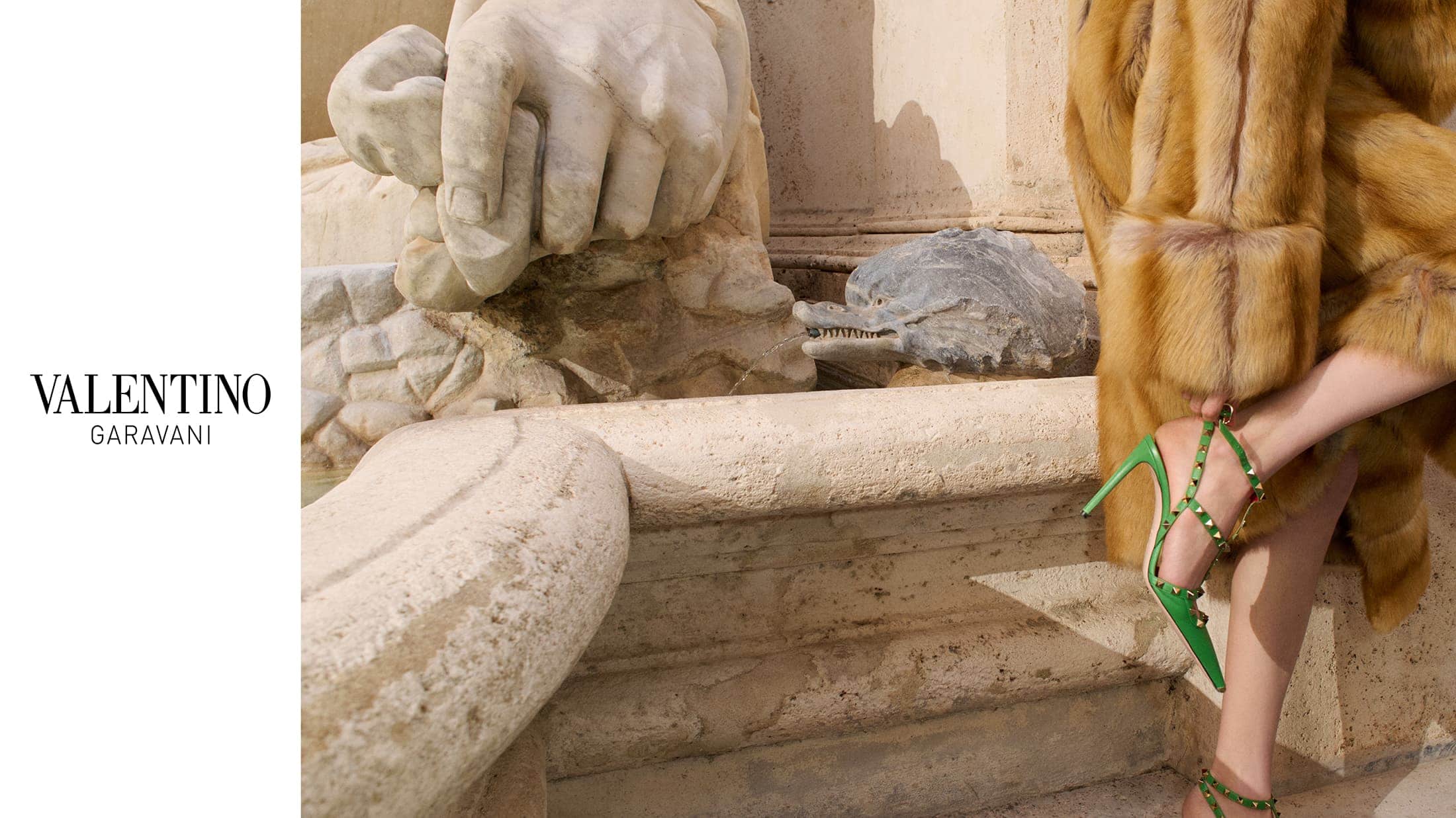 Model wearing green studded heels and a textured fur coat standing near a classical stone fountain.