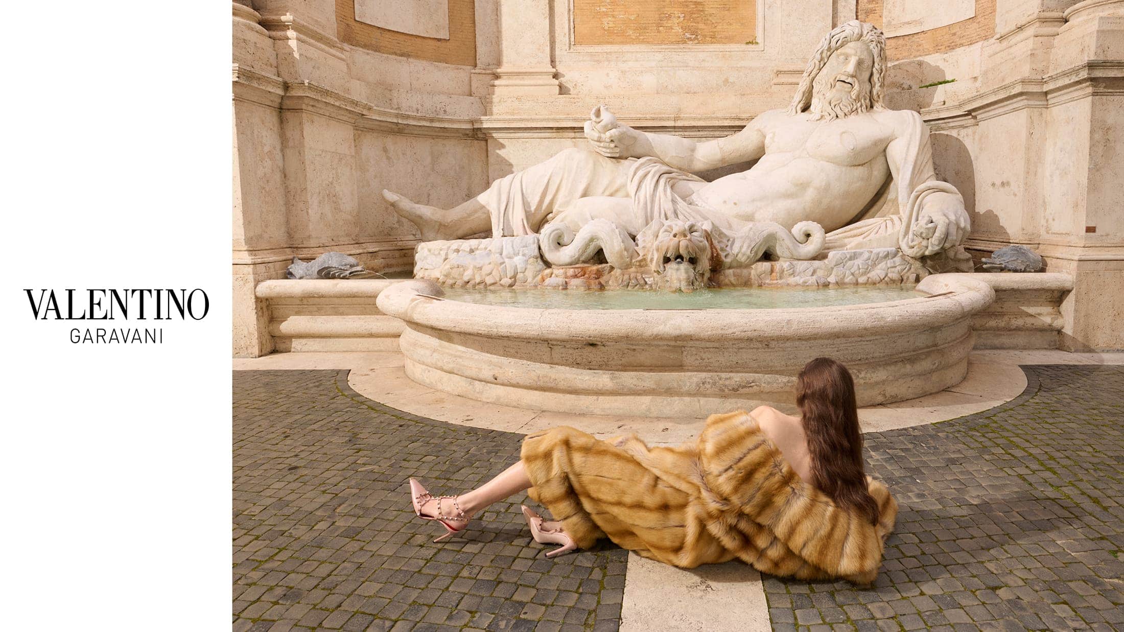 Model lying on cobblestone square wearing a fur coat and rockstud heels beneath a stone fountain sculpture