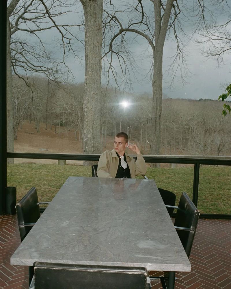 An adult male sits at an outdoor table with a woodland view, wearing a tan jacket over a black shirt in natural daylight.
