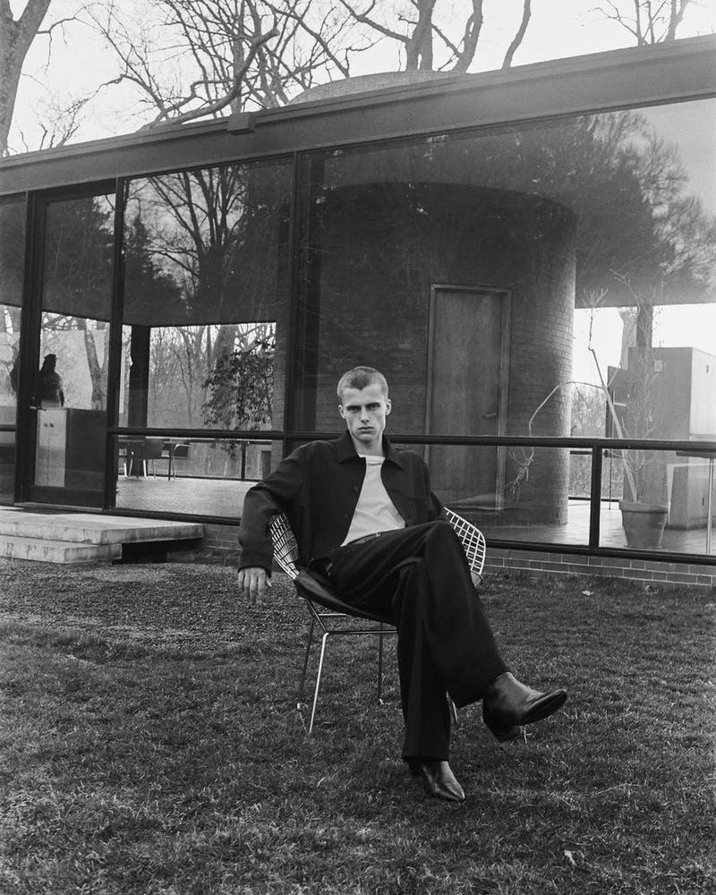 Monochrome portrait of a stylish adult male sitting in a wire chair on a lawn in front of a glass-walled modern house.