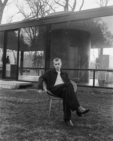 Monochrome portrait of a stylish adult male sitting in a wire chair on a lawn in front of a glass-walled modern house.