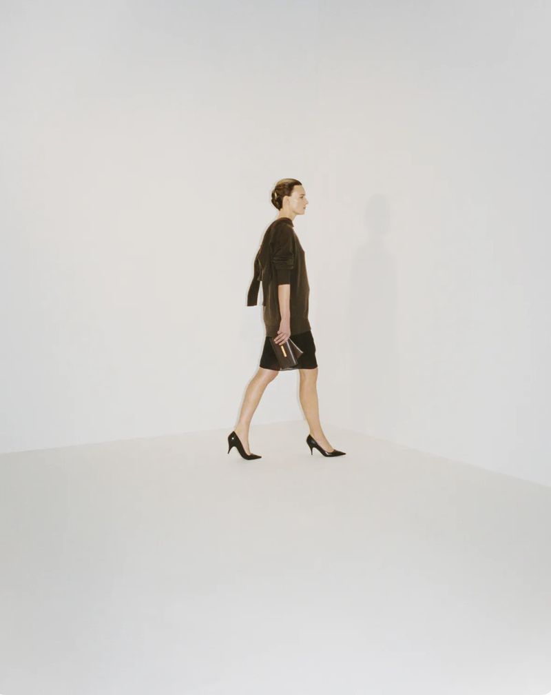 Model walking in profile wearing a dark tunic and skirt against a seamless white studio background