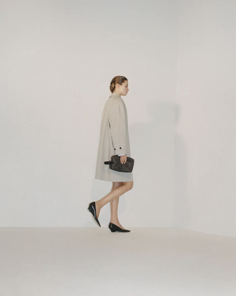 Model walking in profile wearing a neutral beige coat and carrying a dark pouch against a white studio background