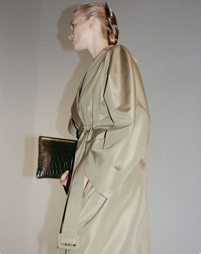 Profile view of a woman wearing a tied belted coat holding a dark structured clutch against a neutral wall