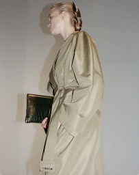 Profile view of a woman wearing a tied belted coat holding a dark structured clutch against a neutral wall