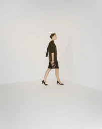 Model walking in profile wearing a dark tunic and skirt against a seamless white studio background