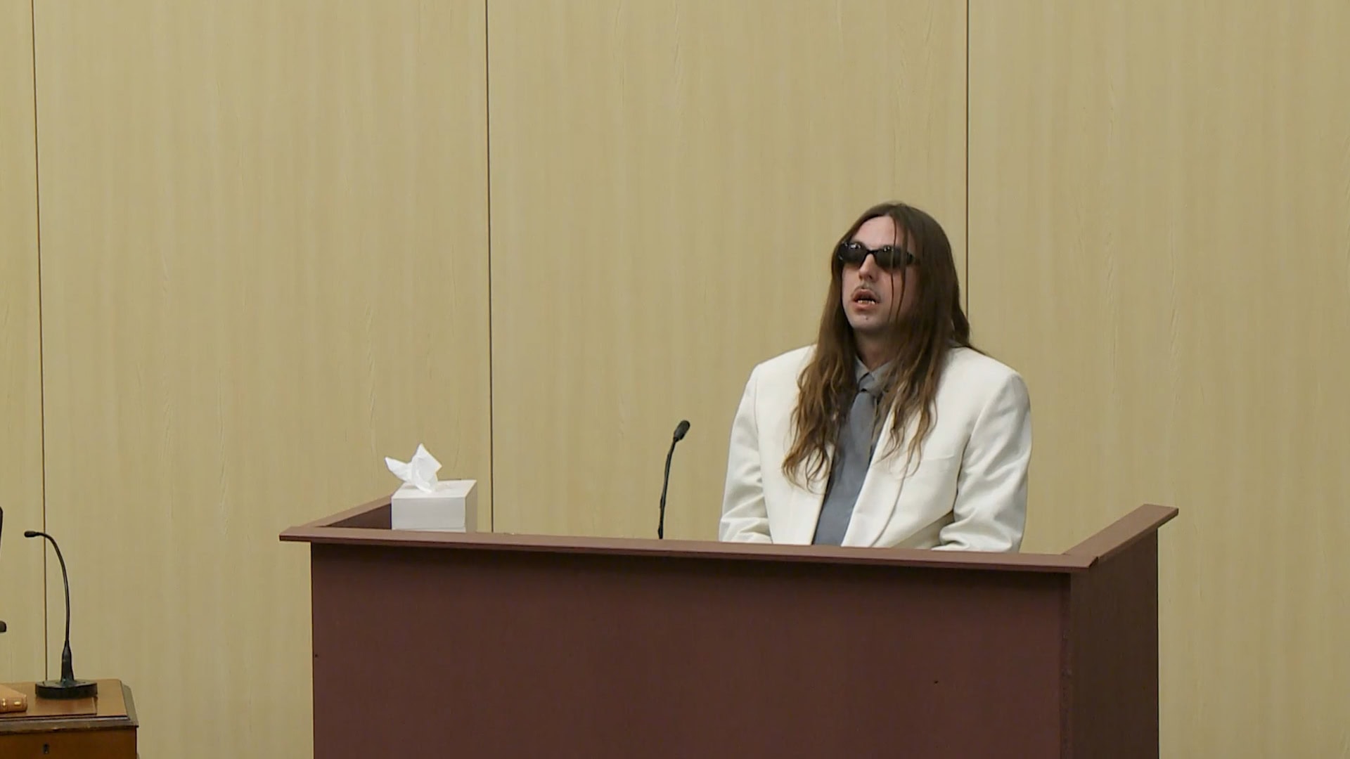 Long-haired person in a white suit and sunglasses standing behind a brown witness stand for Chimi ad campaign.