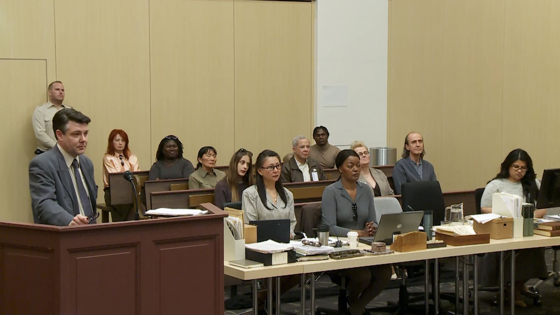 A man in a grey suit standing at a wooden podium inside a courtroom filled with seated people for The Trial ad campaign by Chimi