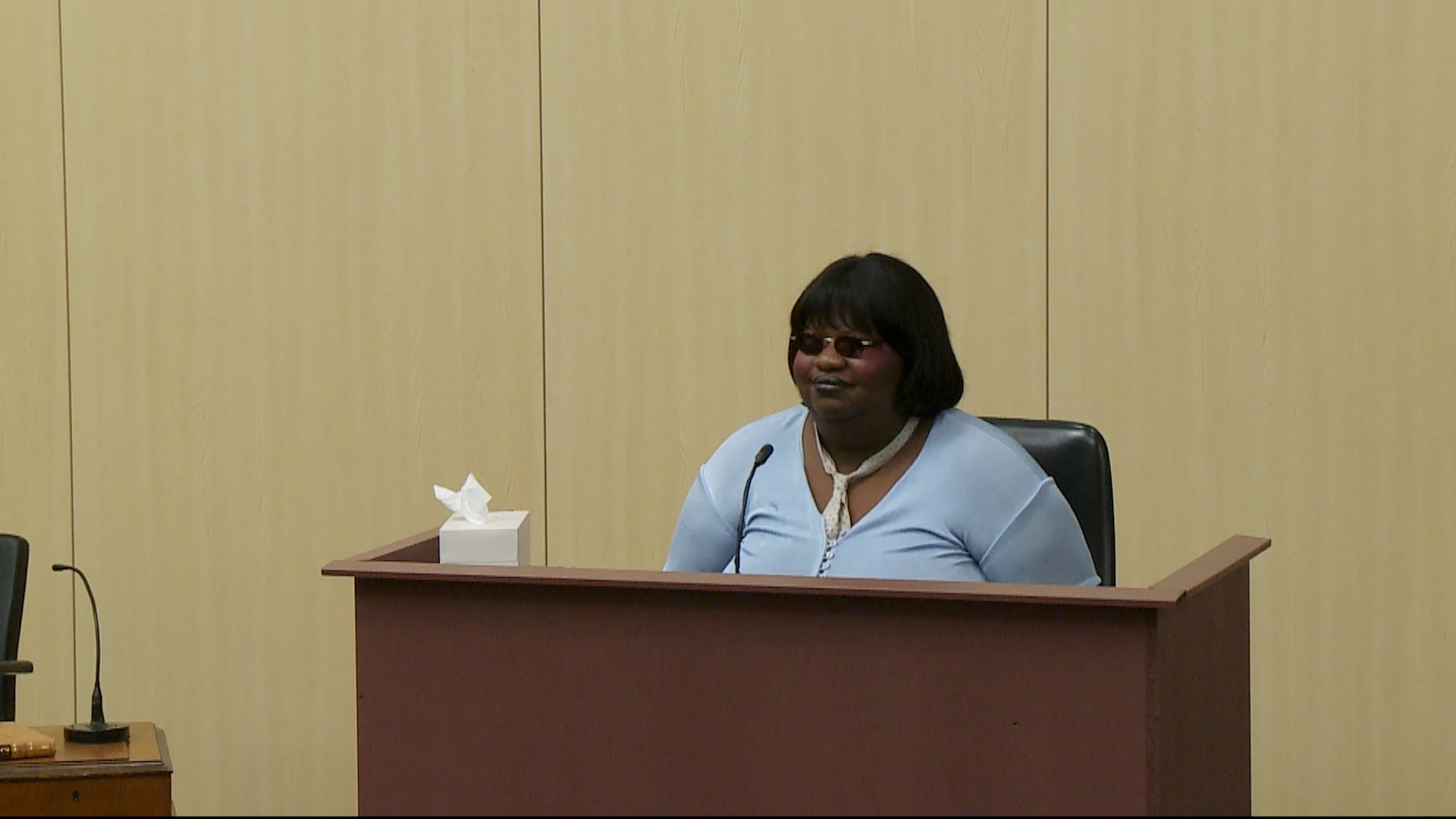 A woman wearing dark sunglasses and a light blue top sitting behind a witness stand in a courtroom.