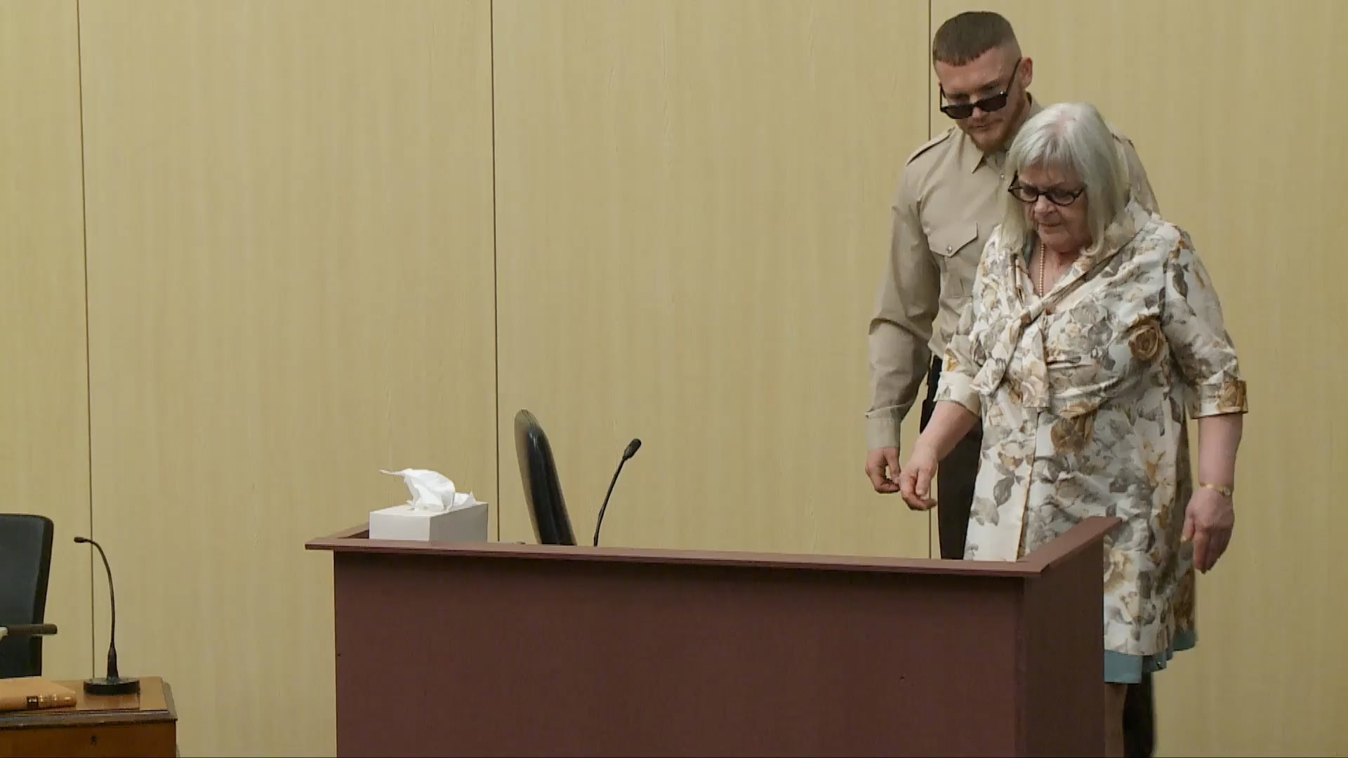 Elderly woman in floral dress walking past a wooden podium with a security guard standing behind her