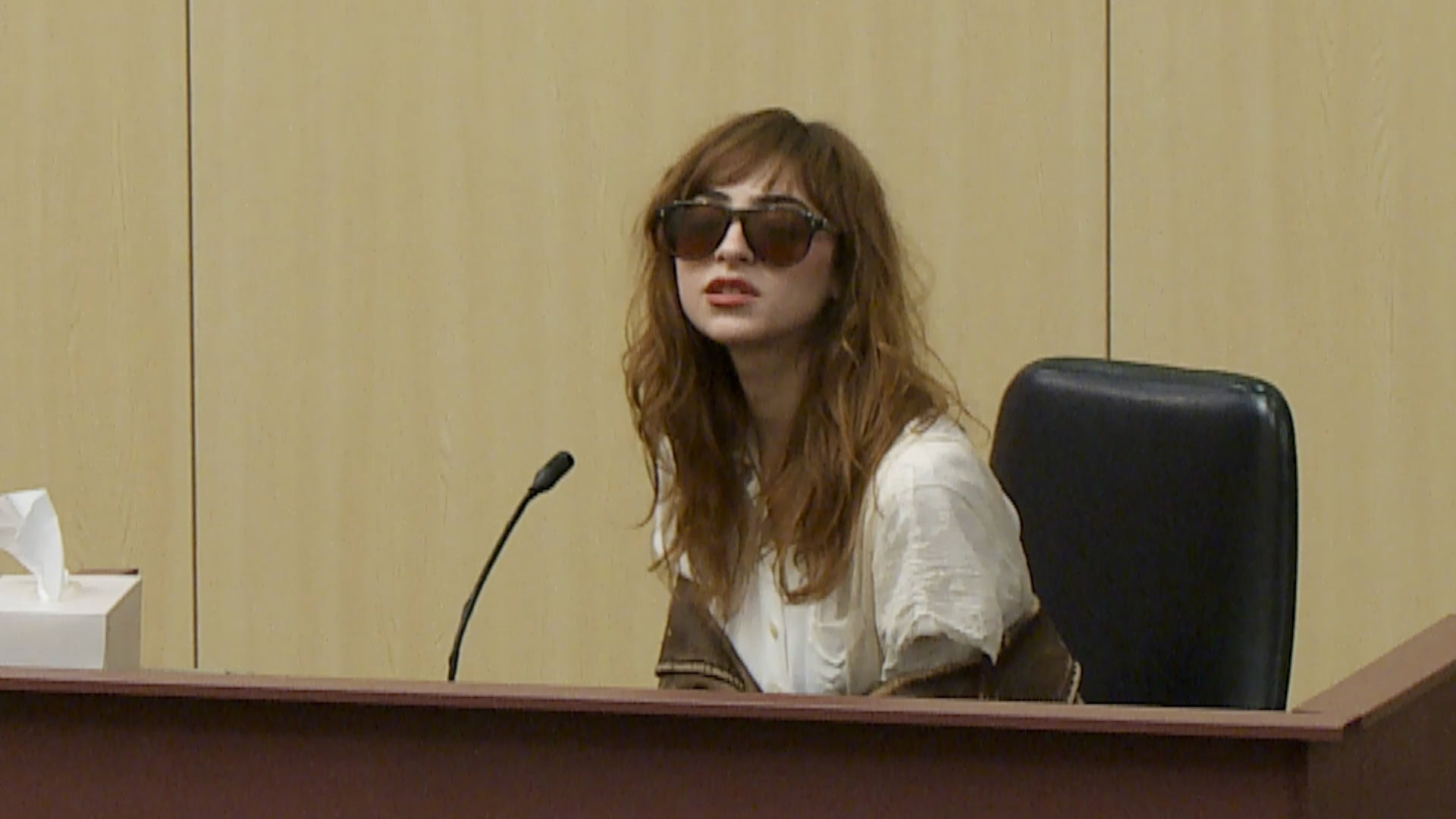 Woman in sunglasses and white shirt seated at a wooden desk with a microphone, in a courtroom setting.