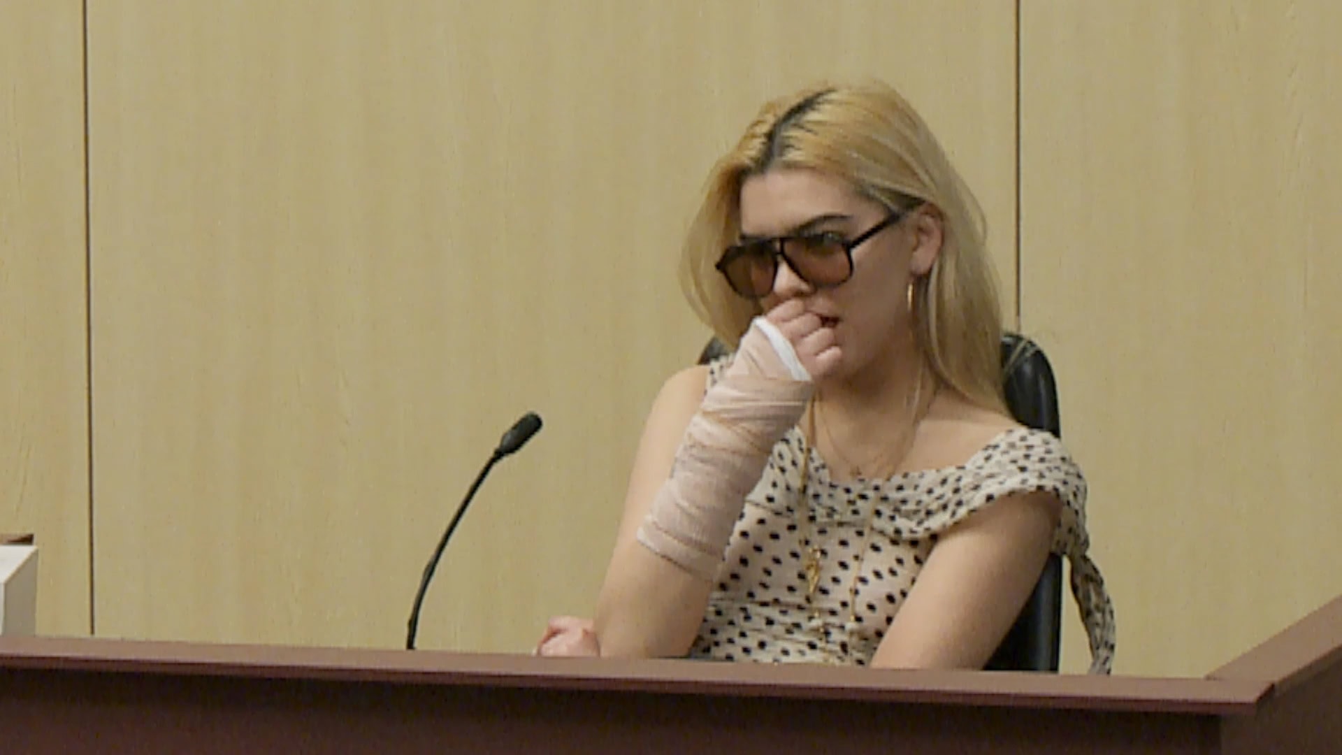 Woman in dark sunglasses and spotted top with a bandaged arm seated behind a wooden desk with a microphone