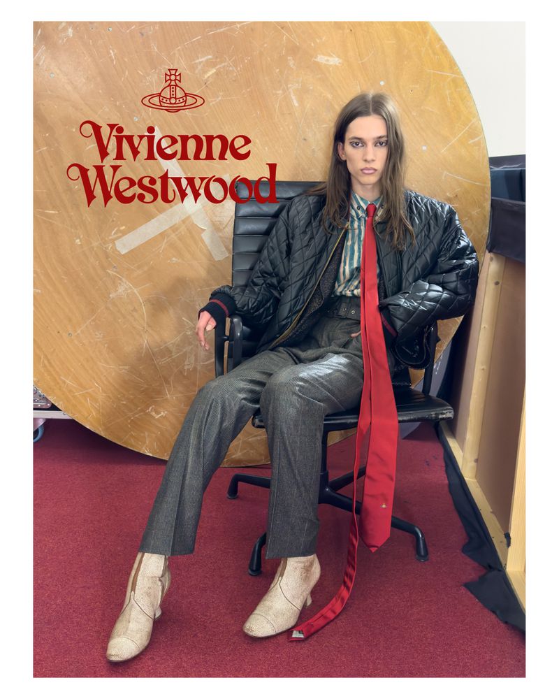 A person wearing a black quilted jacket and grey pants sits on a black office chair in front of a wooden panel with Vivienne Westwood text.