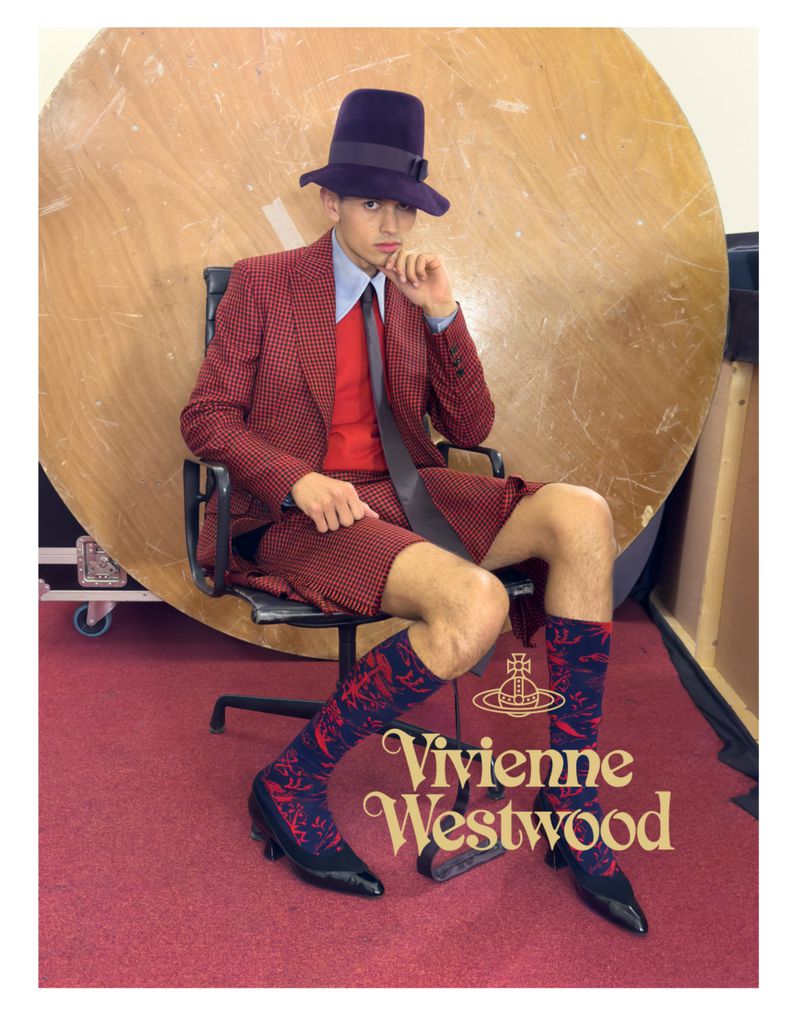 Fashionable young man sits in a chair wearing a red suit with shorts, patterned socks, black heeled shoes, and a distinctive purple hat.