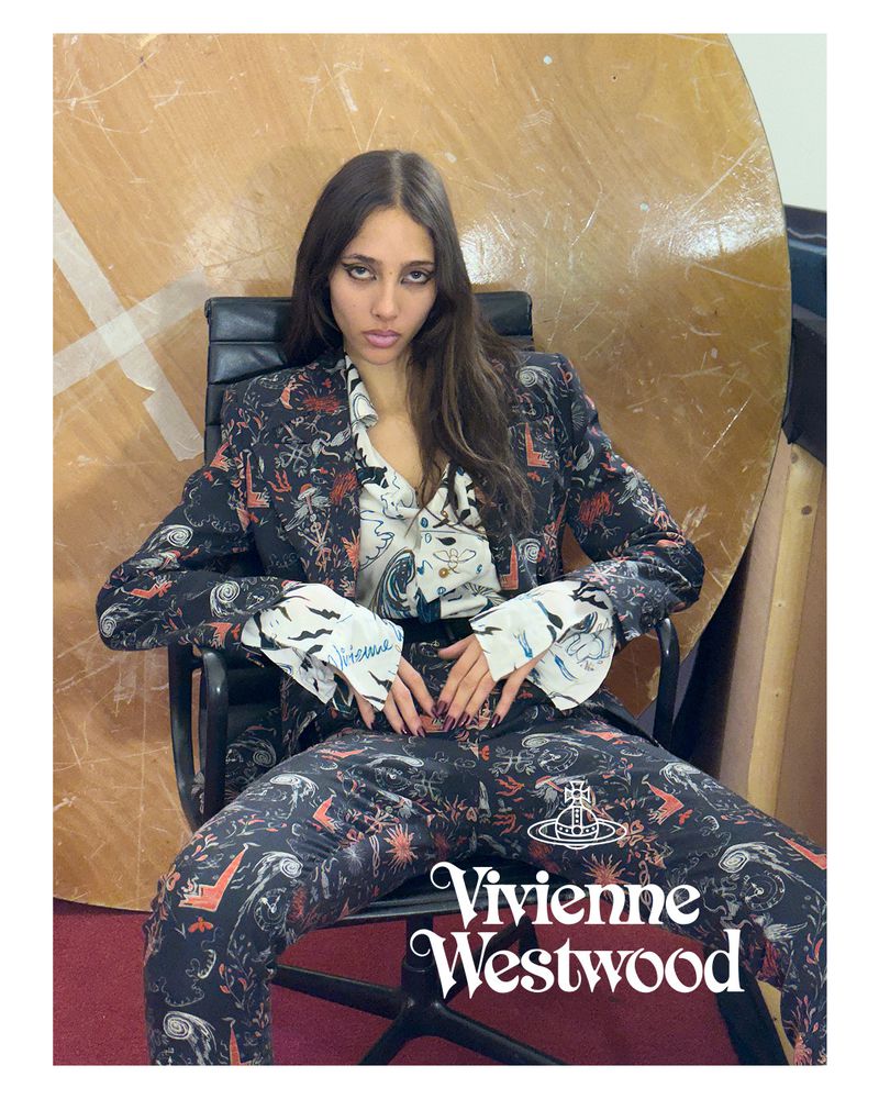 A young adult fashion model poses in a patterned pantsuit and blouse, seated on a black chair against a studio backdrop.