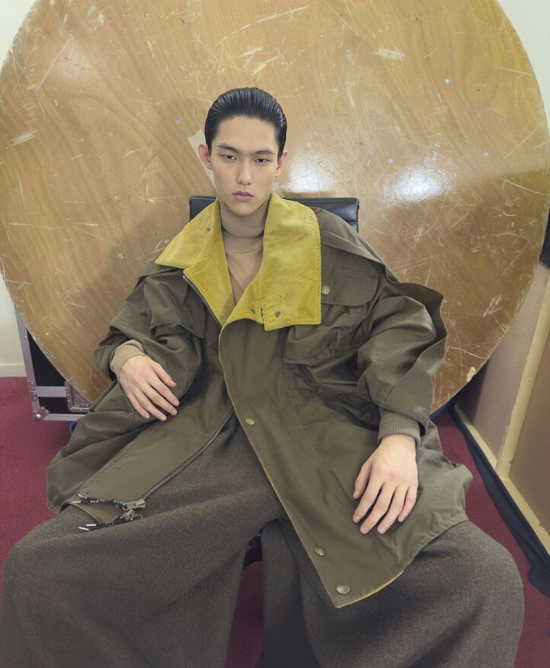 Asian male fashion model in a green oversized jacket and brown wide-legged trousers sitting indoors against a round backdrop.