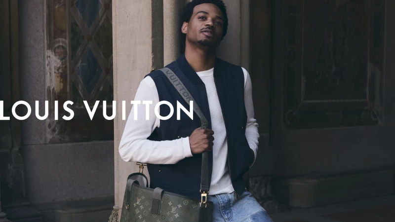 Tyshawn Jones leaning against a stone pillar wearing a navy vest over a white sweater with a monogram shoulder bag for Louis Vuitton