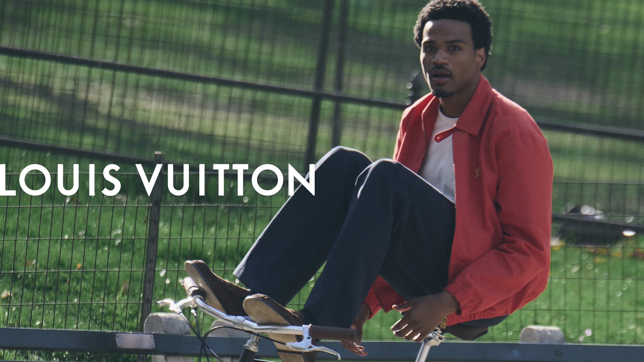 Black man wearing a red jacket and dark trousers riding a bicycle outdoors near a park fence for Louis Vuitton