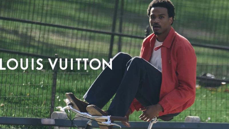 Black man wearing a red jacket and dark trousers riding a bicycle outdoors near a park fence for Louis Vuitton