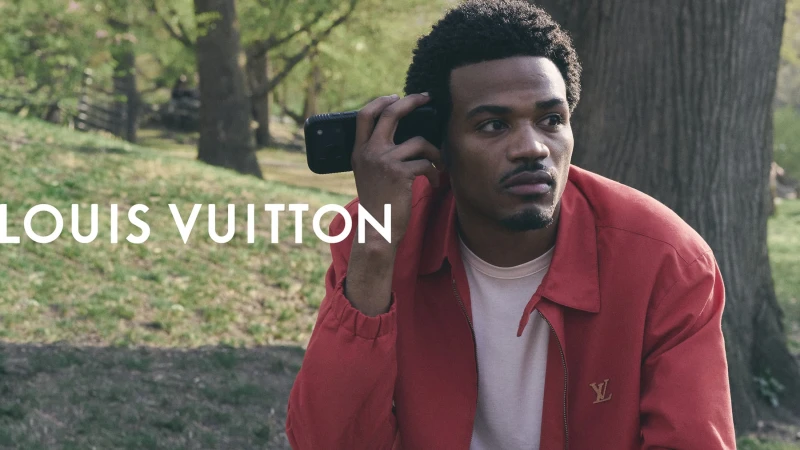 Man wearing a red jacket holding a smartphone to his ear outdoors in a park setting for Louis Vuitton Pre-Fall 2026