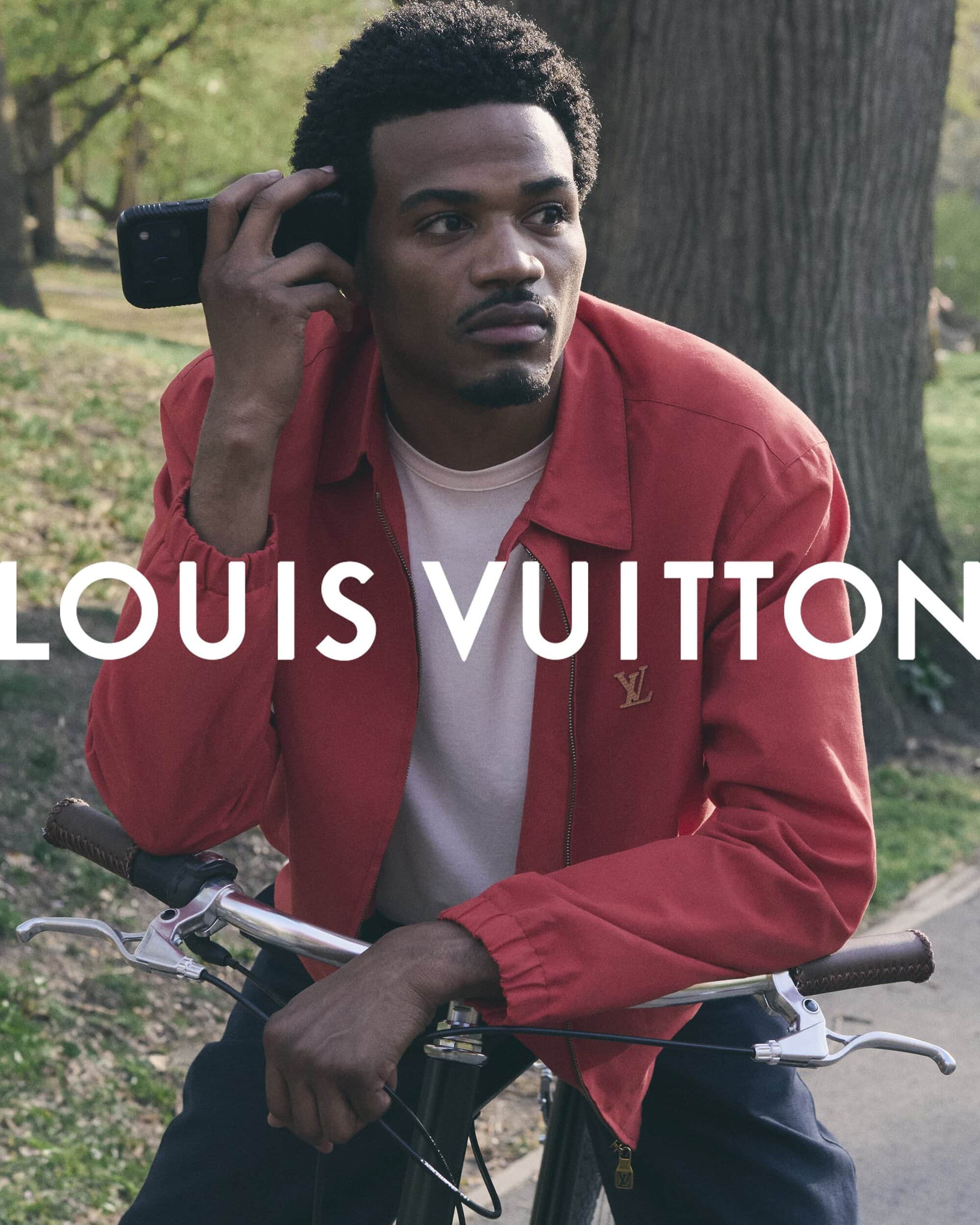 Black man in a red jacket leaning on a bicycle in a park holding phone to ear