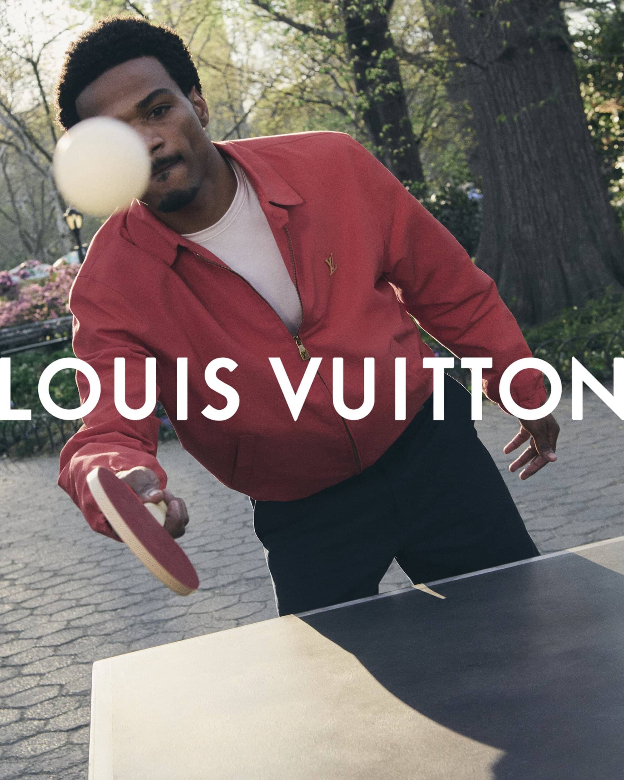 Man playing table tennis outdoors wearing a red jacket and holding a paddle near a table in a park
