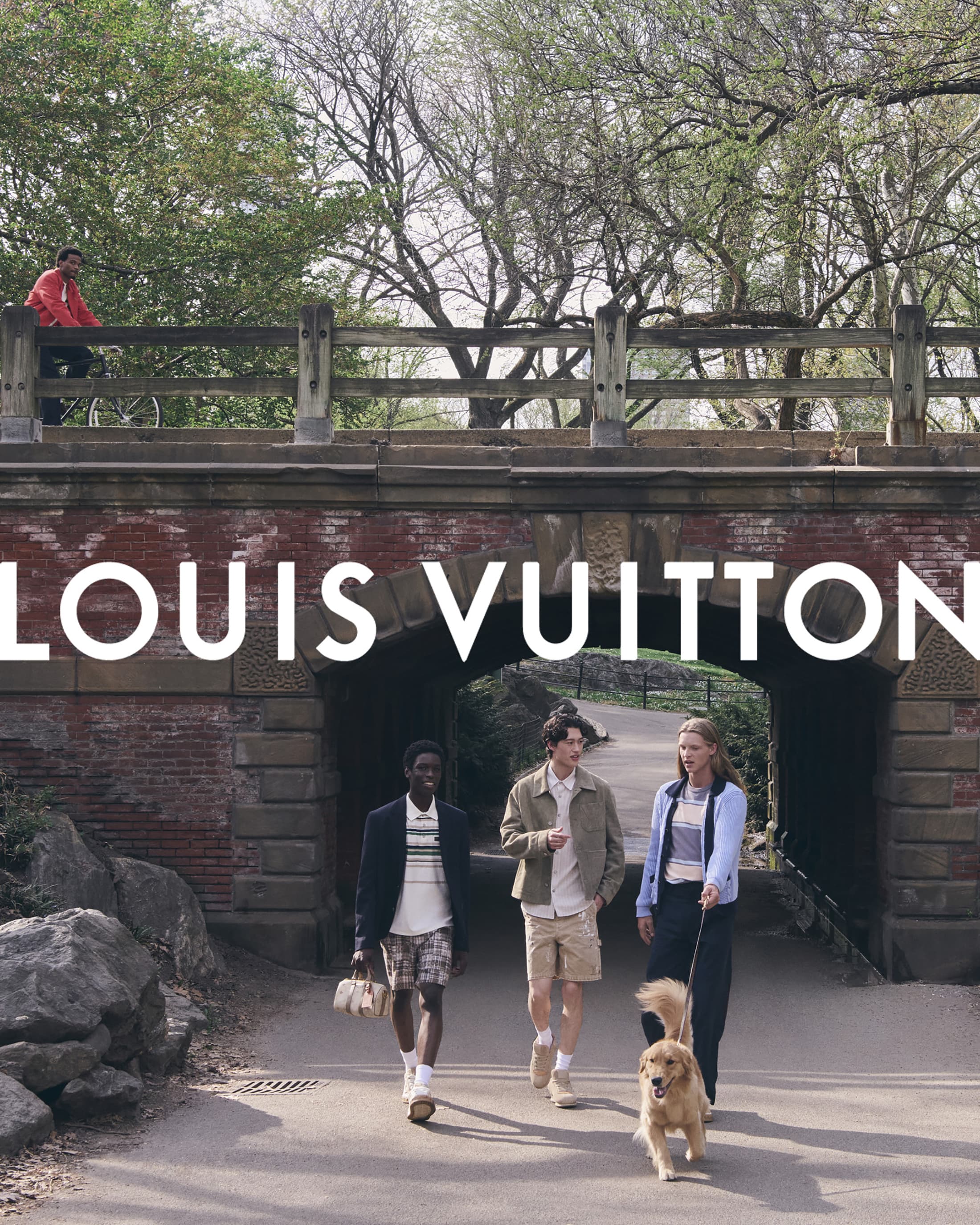 Three models walking together under a brick bridge in a park, with a dog on a leash and a cyclist on the bridge above.