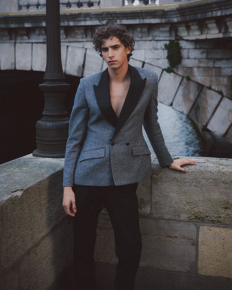Stylish young man in a grey suit with black lapels poses confidently outdoors on a stone structure with a blurred river background.