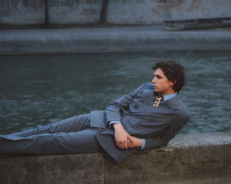 Male model in a grey double-breasted suit reclining on a stone wall beside water for GQ Turkey