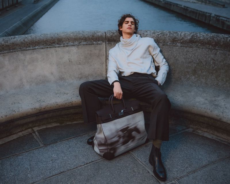 Male model seated on a stone ledge outdoors holding a large patterned leather travel bag for GQ Turkey editorial