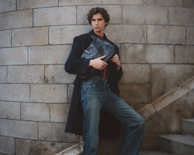 Male model wearing a dark coat and blue jeans holding a dark leather bag against a stone wall for GQ Turkey.
