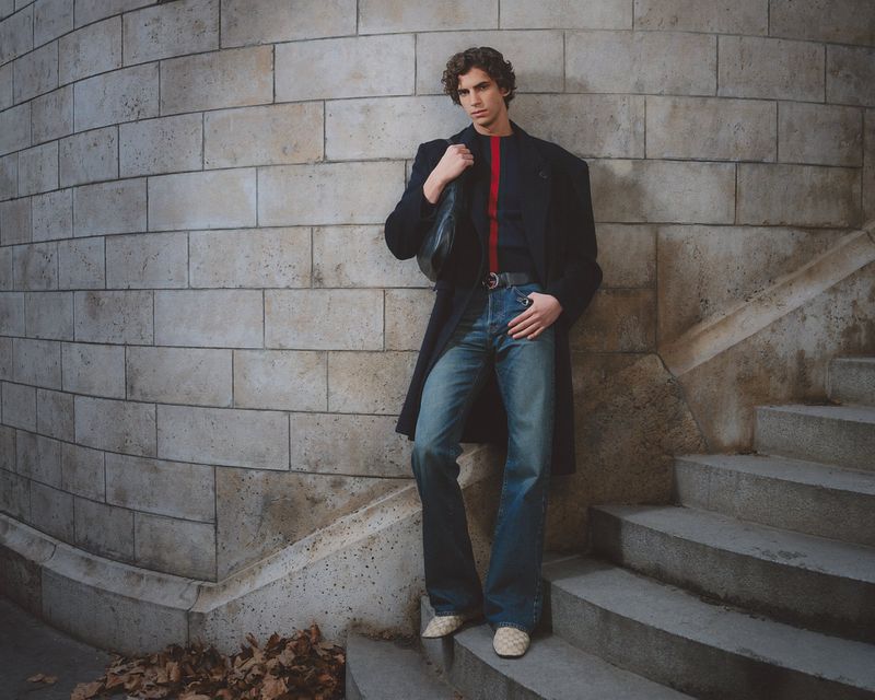 Male model wearing black overcoat and blue jeans standing against curved stone brick wall by staircase for GQ Turkey.