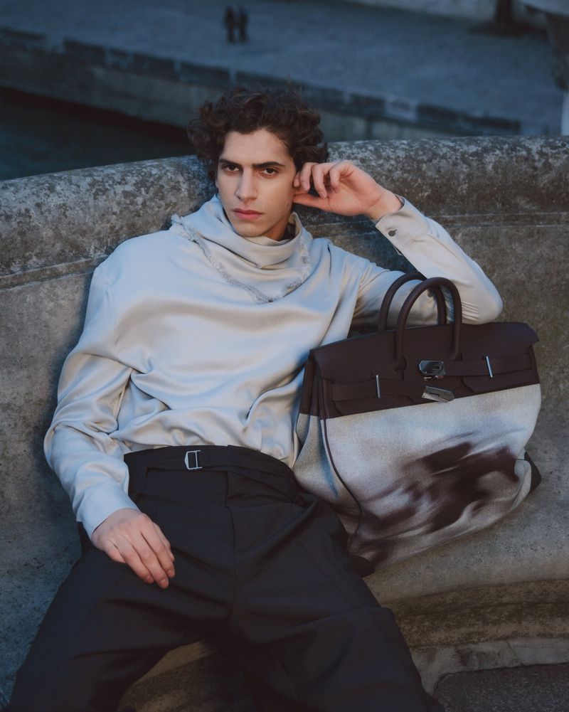 Male model seated on stone ledge leaning against a large luxury leather handbag for GQ Turkey editorial