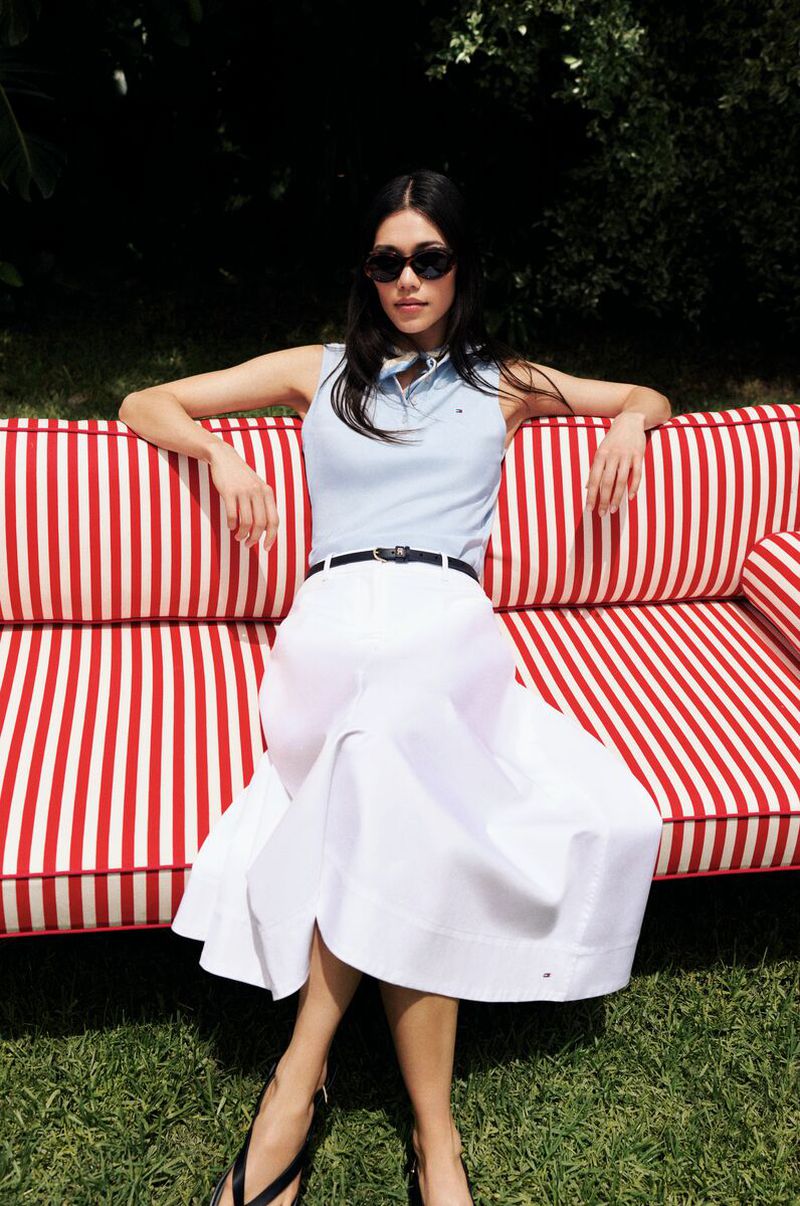 A woman models wearing a light blue polo shirt and white skirt while sitting on a red and white striped couch in a verdant outdoor setting.