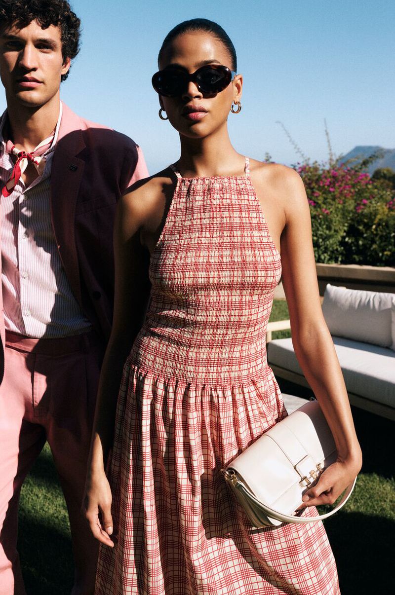Woman in red patterned halter dress holding white handbag standing beside man outdoors in sunny light