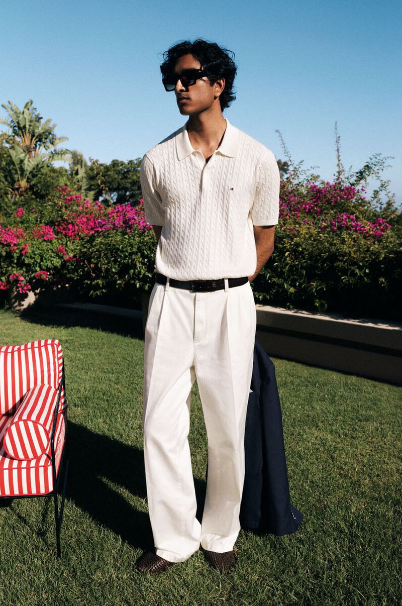 A young man in a white polo shirt and white pants stands outside on the lawn, posing in front of a flower bush under a clear sky.
