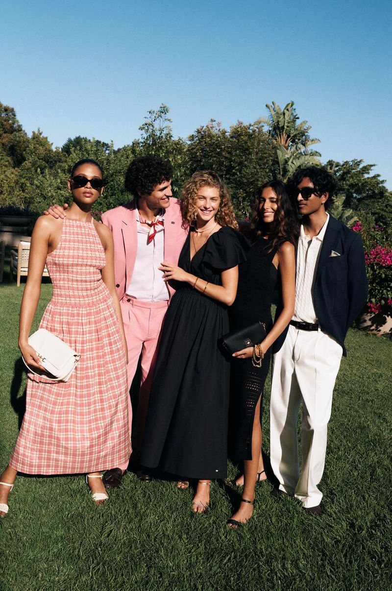 Group of five fashionably dressed adults pose on a grassy lawn in a sunny outdoor setting; various dresses, suits, and accessories.
