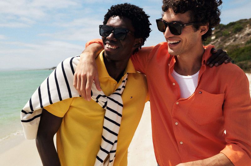Two young men smile on a sunny beach; one wears a yellow polo with a striped sweater, the other a coral shirt and sunglasses.