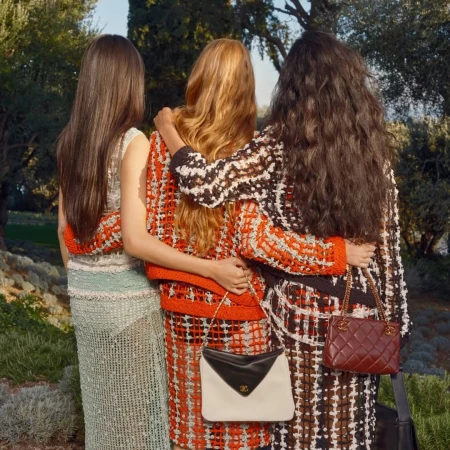 Three female models with long hair from behind wearing tweed skirts and jackets with chain handbags in an outdoor garden.
