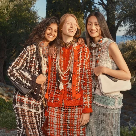 Three female models posing outdoors in textured luxury fashion garments against a background of trees and coastal blue water.